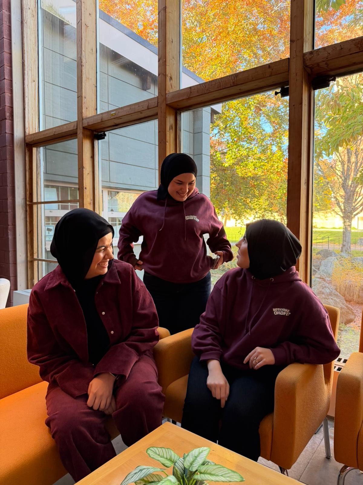 Tre medarbetare i vinröda Ordfest-hoodies skrattar tillsammans i en ljus lokal med höstlöv utanför fönstren.